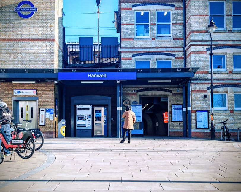 A woman standing outside the entrance of Hanwell station, part of the Elizabeth Line in London, with her back facing the camera. She is wearing a light brown coat, dark pants, and sneakers. The statio