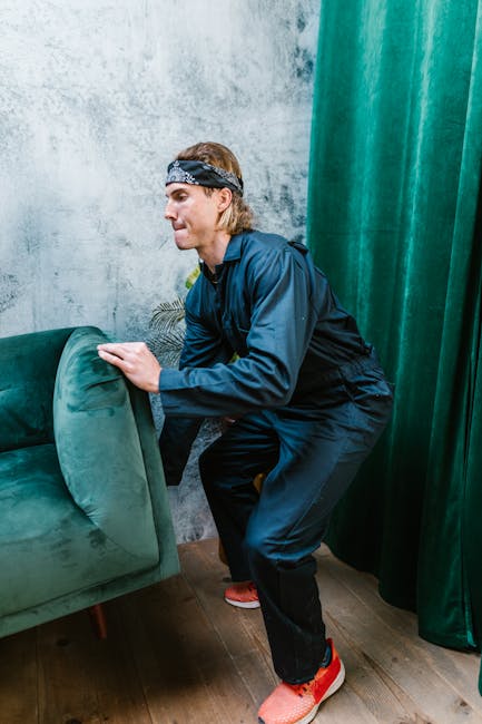 A person with shoulder-length hair, wearing a black headband, navy blue jacket, and black trousers, is shown in an indoor setting, lifting a green upholstered armchair with wooden legs. The individual is positioned in front of a concrete-textured wall and next to dark green curtains, suggesting a room prepared for moving or packing. The person appears focused on the task, using both hands to grip the chair, which is part of the furniture being loaded into a van or transported within a home during a furniture move. The scene highlights the careful handling of a piece of soft furniture in a home relocation process, with the environment illuminated by natural or ambient indoor lighting, indicating a professional move or packing operation by Man With a Van Ealing, as part of house removals or furniture transport services.