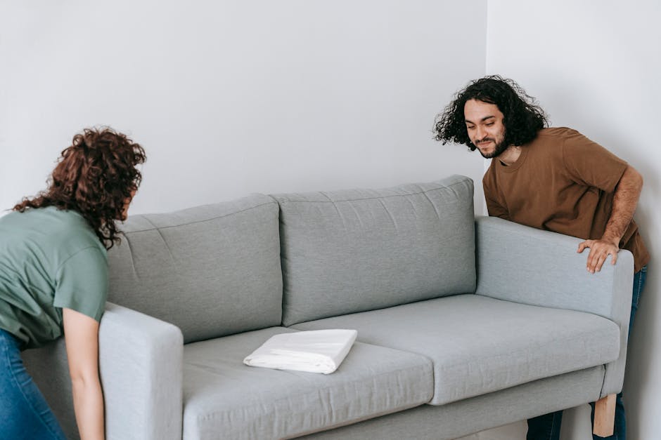 Two individuals are engaged in a home relocation process inside a room, preparing a grey fabric sofa for moving. The person on the left, with curly brown hair and wearing a green shirt, is in the process of lifting or stabilizing the sofa from the front end, while the person on the right, with curly dark hair and a brown shirt, is holding the armrest on the opposite side, supporting the loading process. The sofa is positioned against a plain white wall, with a neatly folded white blanket resting on the middle cushion, indicating a careful packing and handling effort. The lighting in the room appears natural, highlighting the focused actions of the movers during the furniture transport. In the background, the setting provides a neutral environment typical for a residential home, and the scene exemplifies the proper techniques used by professional removals services such as Man With a Van Ealing to facilitate furniture transport amidst home relocation tasks.