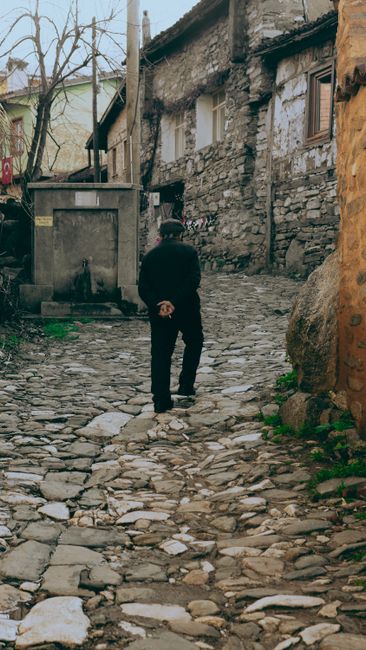 A man dressed in dark clothing stands on a narrow, cobbled street in a residential area, with his back to the camera and hands clasped behind him. The street is lined with old, weathered buildings constructed from stone and brick, with small windows and wooden shutters. A large, grey metal container is positioned on the left side near the corner of one building, while a patch of green moss and small plants grow at the base of rocks and walls on the right. The scene is lit with natural daylight, highlighting the textured surfaces of the uneven stones and aged facades. This setting appears to be part of a historic or traditional neighbourhood, where home relocation or furniture transport may involve navigating narrow streets and cobbled pathways, a consideration relevant to local removal services such as those offered by Man With a Van Ealing.