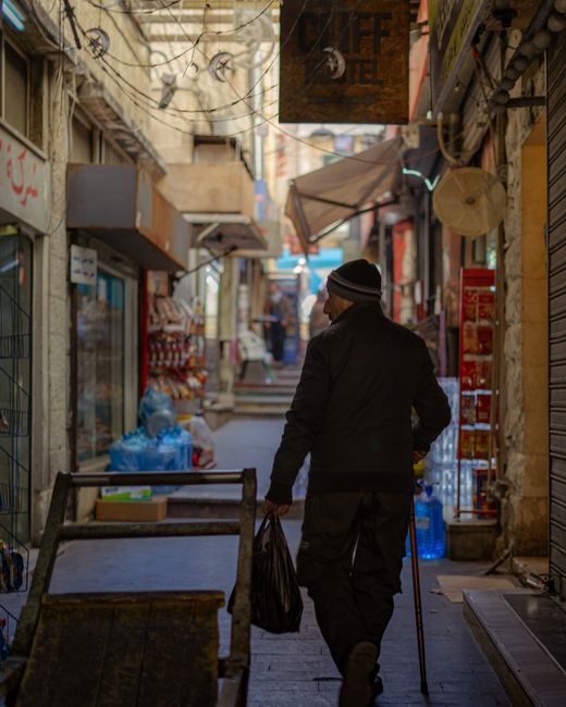 A man wearing a dark jacket, trousers, and a beanie hat is walking through a narrow outdoor street or alleyway, carrying a black plastic bag in his right hand and using a cane in his left hand. The alley is lined with small shops and market stalls, some with metal shutters partially closed, and window displays with various goods. Overhead, there are string lights and hanging signs, including one wooden sign reading 'OFF HOTEL,' and a beige paper lantern. The ground is paved with cobblestones, and waste bags and miscellaneous items are visible near the shop entrances. The scene appears to be in an urban setting, possibly during a busy day, with a mix of natural and artificial lighting. Man With a Van Ealing provides services related to home relocation and furniture transport, as suggested by the context of moving-related visuals.