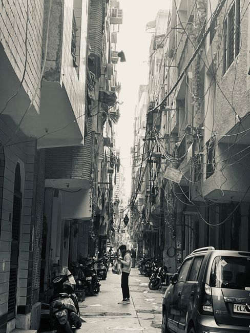 A man dressed in dark clothing stands on a narrow, cobbled street in a residential area, with his back to the camera and hands clasped behind him. The street is lined with old, weathered buildings constructed from stone and brick, with small windows and wooden shutters. A large, grey metal container is positioned on the left side near the corner of one building, while a patch of green moss and small plants grow at the base of rocks and walls on the right. The scene is lit with natural daylight, highlighting the textured surfaces of the uneven stones and aged facades. This setting appears to be part of a historic or traditional neighbourhood, where home relocation or furniture transport may involve navigating narrow streets and cobbled pathways, a consideration relevant to local removal services such as those offered by Man With a Van Ealing.