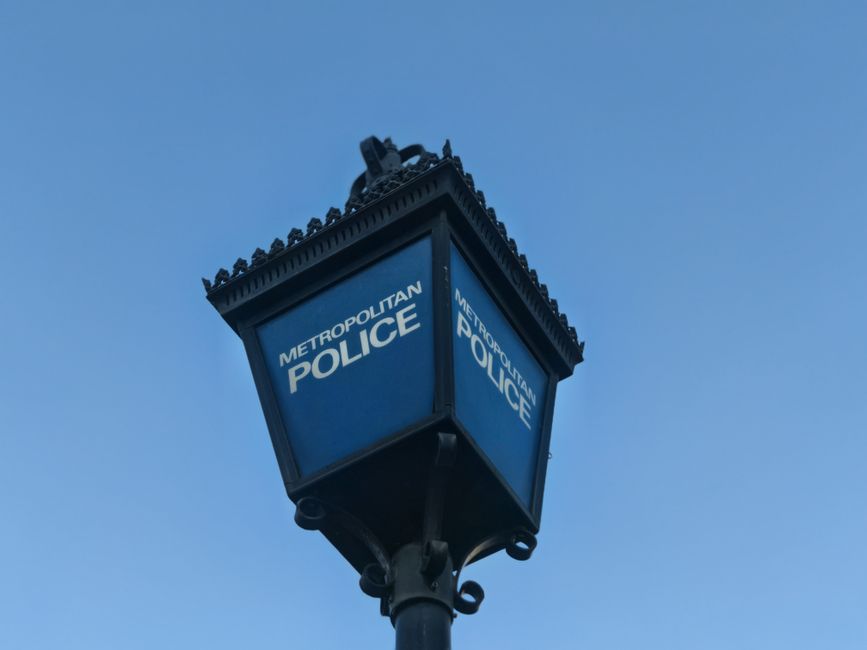 Close-up image of a historic-style street lamp with a black metal post and ornate detailing at the top, mounted outdoors against a clear blue sky. The lamp features a rectangular blue sign with white