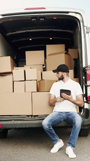 A man wearing glasses and a light blue shirt is seated on a wooden chair just outside the rear of a white delivery van parked on a street in front of a modern building with glass windows. The van's re